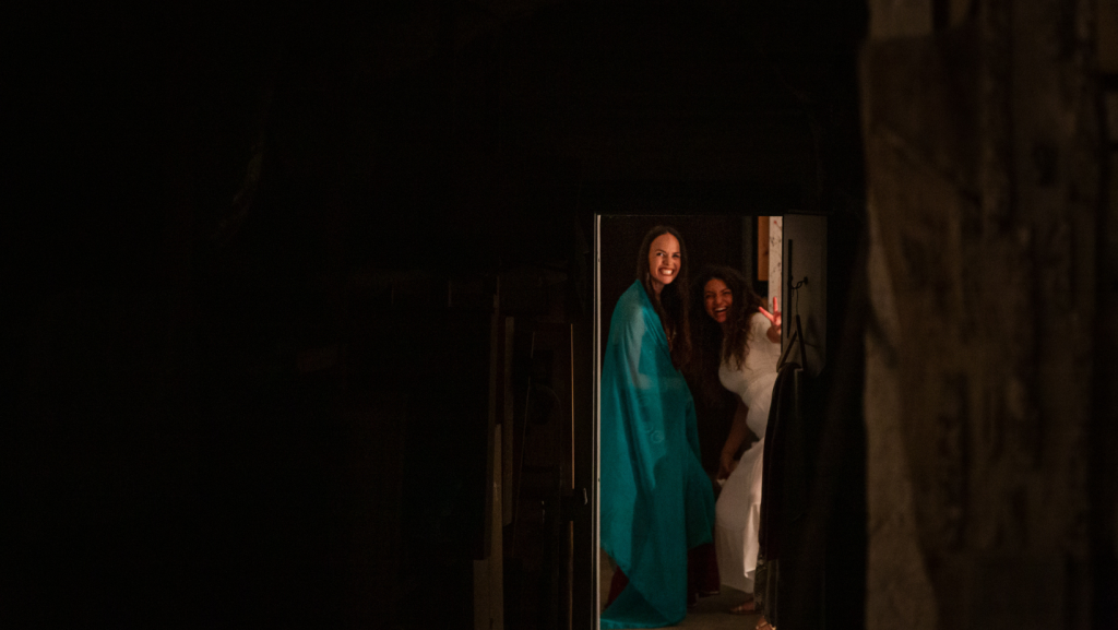 Due donne sorridenti riprese in un momento di spontaneità mentre sbirciano da una porta durante un evento al chiuso.