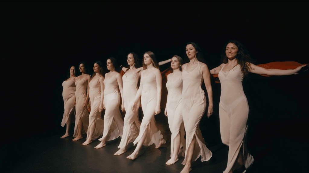 Gruppo di donne in abiti chiari avanza in sincronia sul palco, tenendo un drappo rosso sulle spalle, in una performance coreografica.