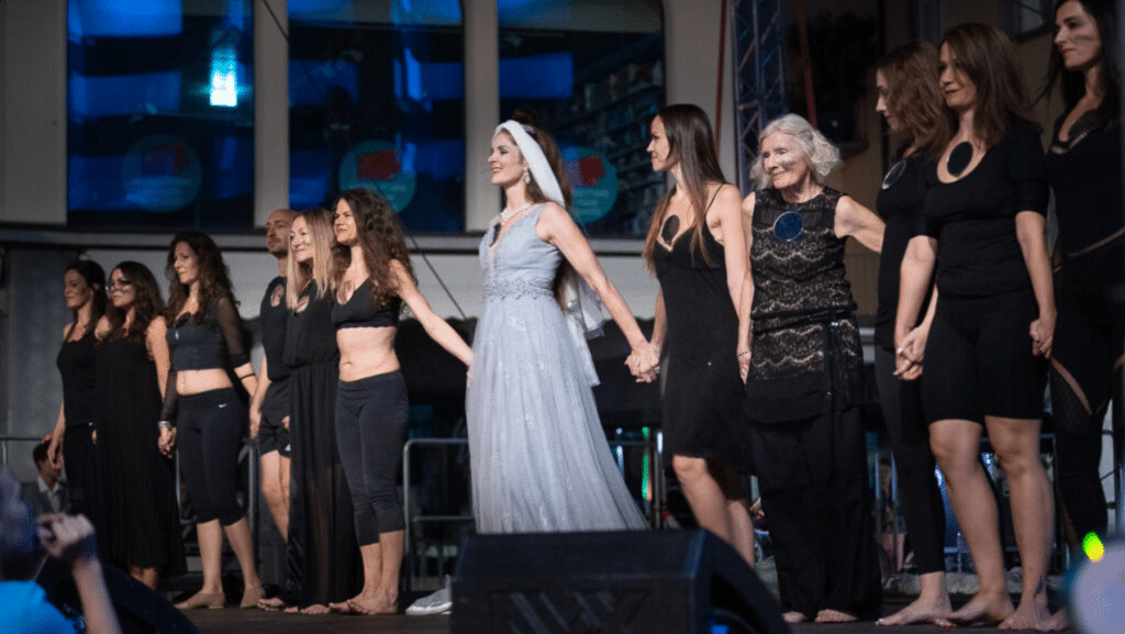 Danzatrici e performer di diverse età si tengono per mano in fila sul palco al termine di una performance.