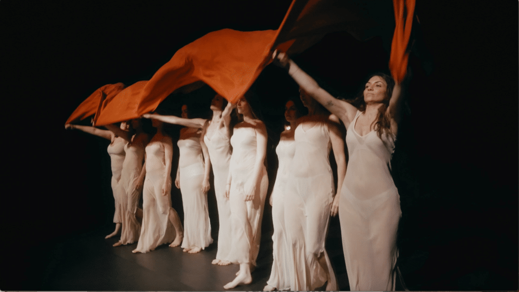Danzatrici in abiti chiari sventolano tessuto rosso durante una performance coreografica in fila sul palco.