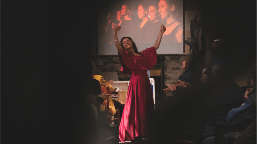 Una performer in abito rosso celebra l'arte e l'educazione, salutando il pubblico durante un evento. Sostienici, il tuo contributo è fondamentale per diffondere progetti artistici ed educativi.